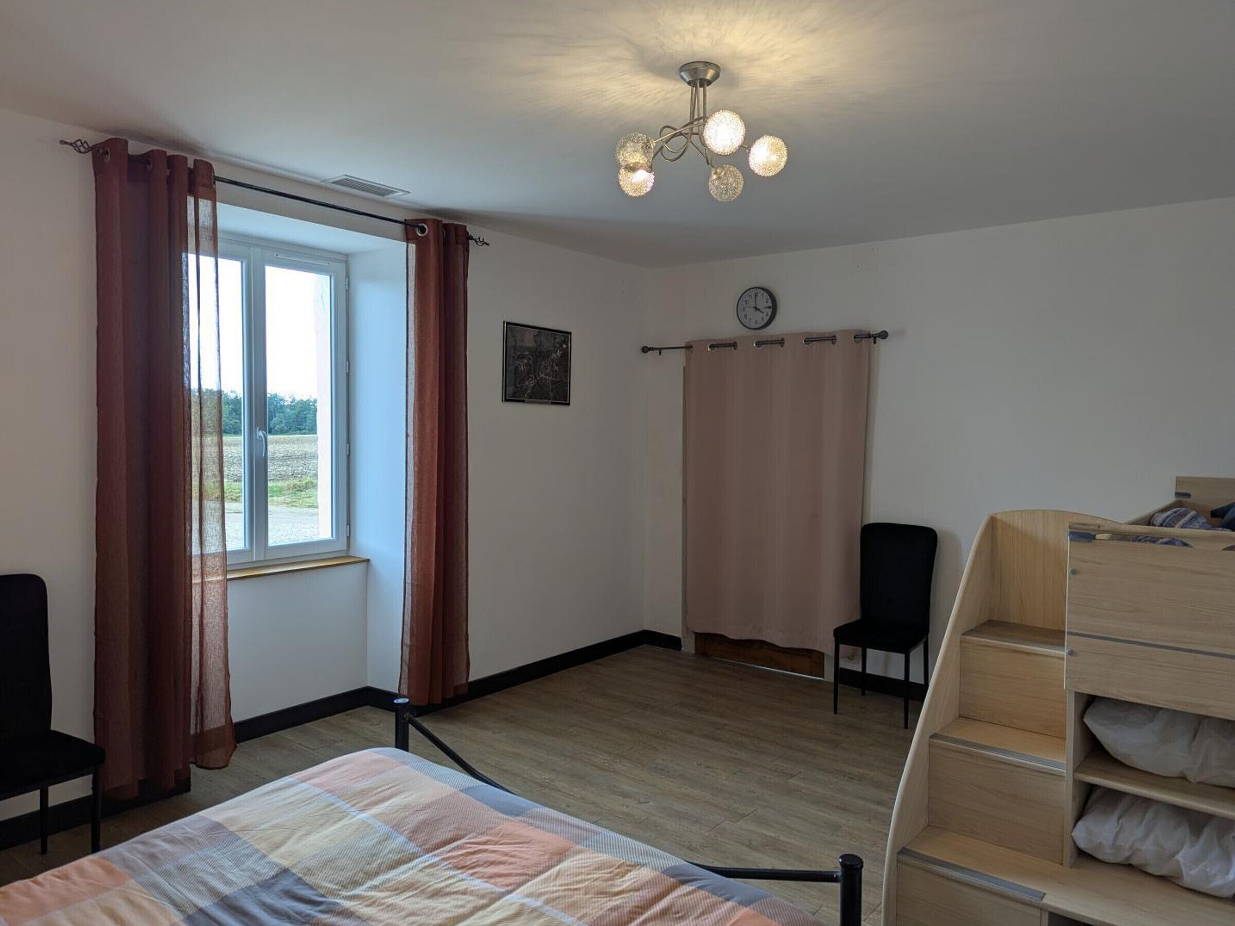 Une chambre à coucher comprenant un lit, une commode en bois, une chaise, une fenêtre avec des rideaux, une horloge et un luminaire au plafond.