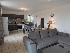 A modern living room with a grey sofa, a dining area with a table and chairs, a kitchen with dark cabinets and a tiled backsplash, and a wall-mounted clock.