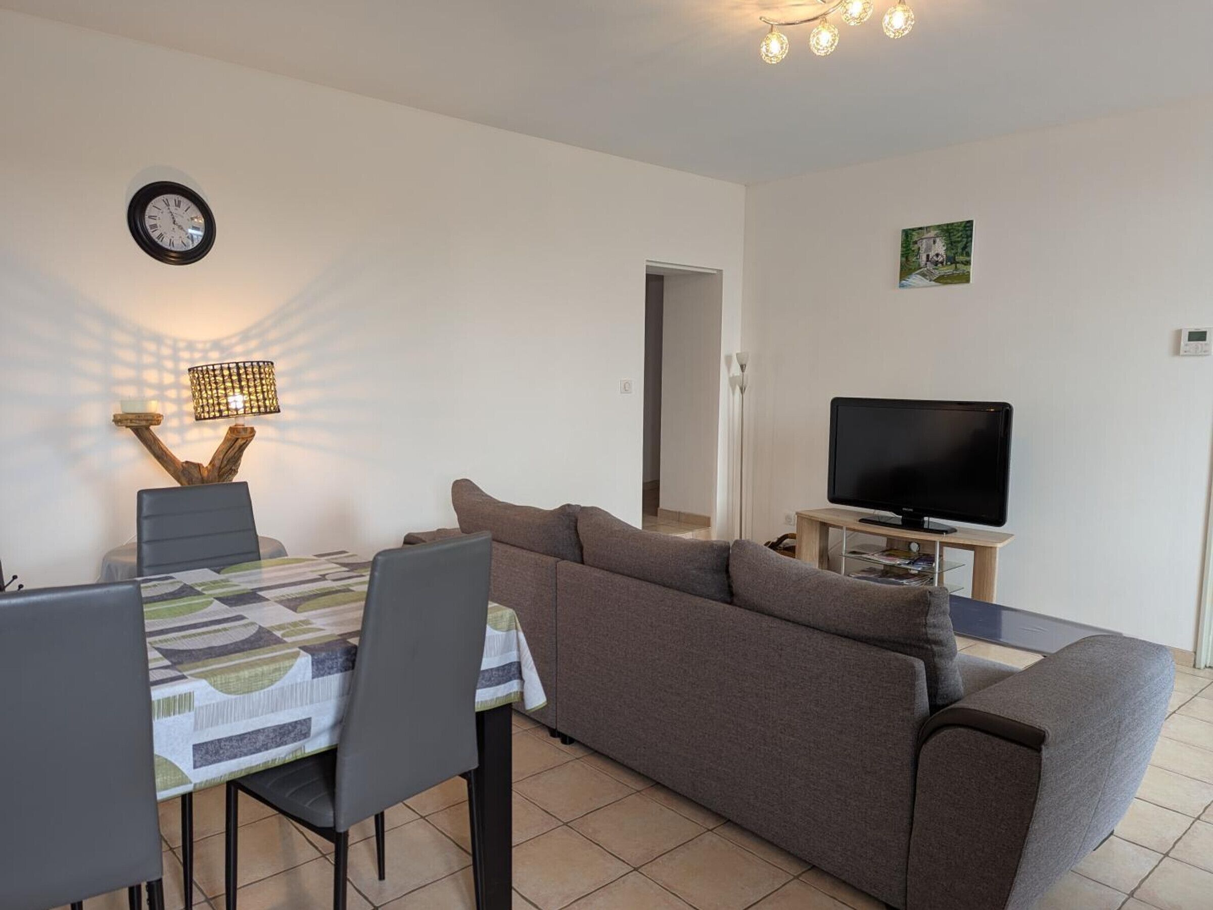 A living room with a sofa, TV, dining table, chairs, lamp, clock, and a door leading to another room.