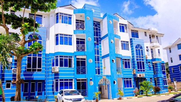 A multi-story building with blue and white exterior walls, large windows, and a paved entrance area with parked cars.