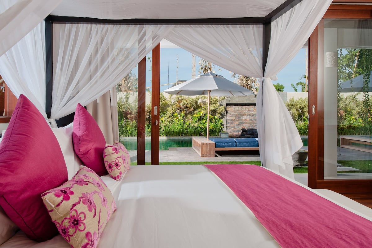 A bedroom with a canopy bed, a view of the garden, and a seating area with an umbrella.