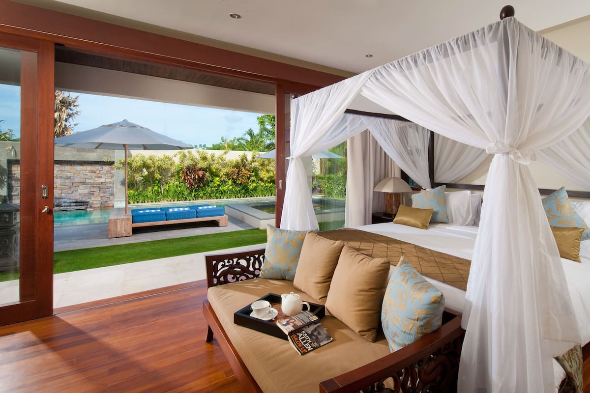 A bedroom with a canopy bed, a sofa with a tray table, and a view of a pool and outdoor seating area.