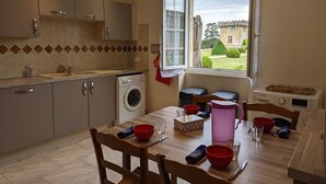 A compact kitchen with a washing machine, a dining table with chairs, and a view of a building through a window. - Charming Cottage with Historic Castle Views, 7 Hectares, Bikes, Near Cognac (Cherves-Richemont)