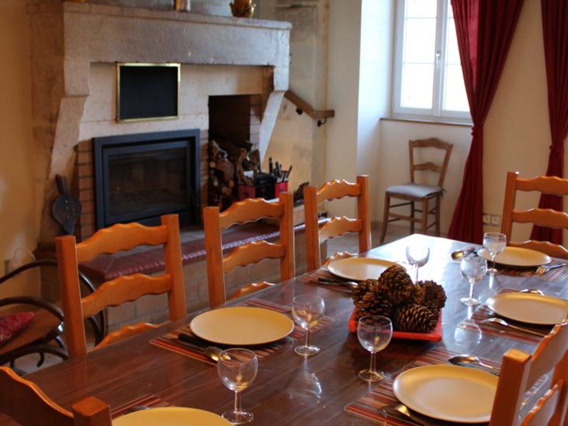 A dining room with a fireplace, wooden chairs, a long table set for four with plates, glasses, and pine cones.