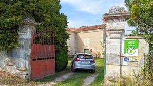 Une porte en pierre surmontée d’une porte rouge, une voiture garée devant, et une enseigne indiquant qu’il s’agit d’un gîte, avec les coordonnées pour prendre contact.