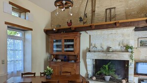 A dining area with a wooden table and chairs, a fireplace with a stone mantle, and a wooden cabinet with glass doors. - Charming stone cottage in Bellon with garden, fireplace and internet (Bellon)