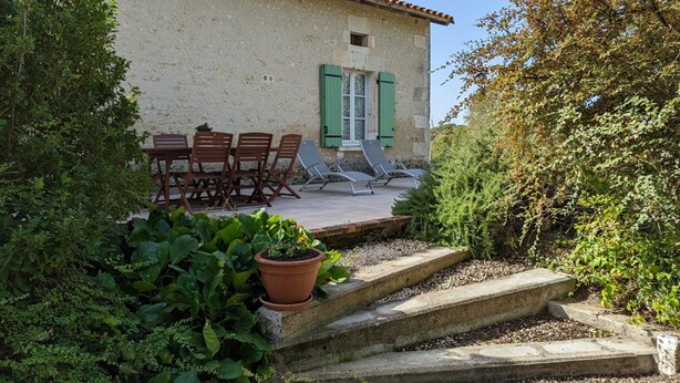A rustic building with a tiled roof, a small window with shutters, and a patio area with a table and chairs. - Charming stone cottage in Bellon with garden, fireplace and internet (Bellon)