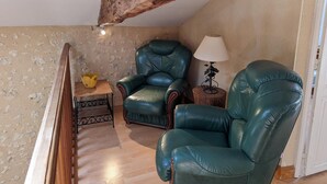 A loft-style room with a high ceiling, exposed wooden beams, two green leather chairs, a small wooden table with a lamp, and a yellow decorative item on a woven stool. - Charming stone cottage in Bellon with garden, fireplace and internet (Bellon)