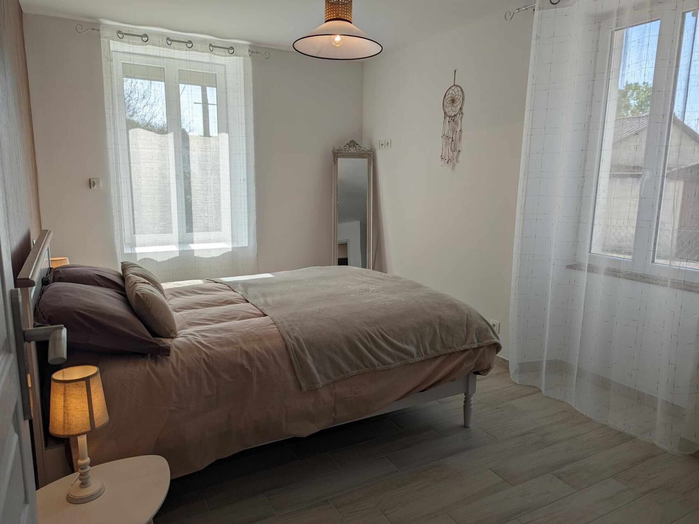 Ein Schlafzimmer mit einem Bett, einem Nachttisch mit Lampe, einem Spiegel und einem Fenster mit Vorhängen.