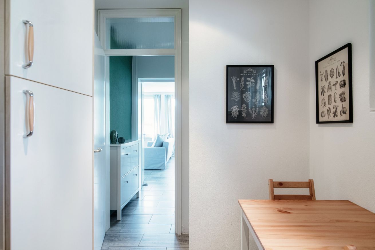 Een gang met een houten tafel en stoelen, een witte kast met lades, en een lijst met een botanische afbeelding aan de muur.