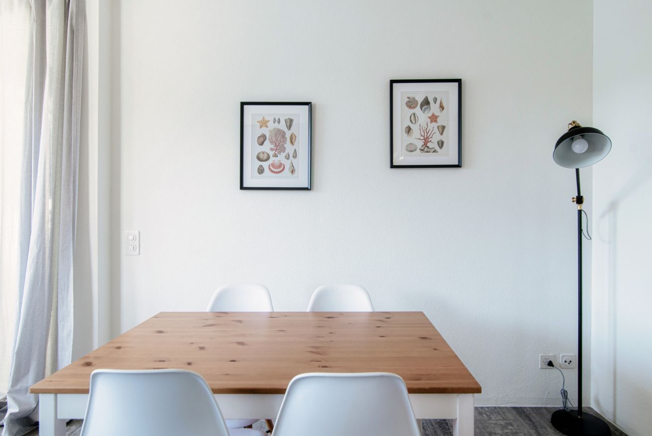Ein Zimmer mit einem Holztisch, weißen Stühlen, zwei gerahmten Bildern an der Wand, einer Stehlampe und einem Fenster mit Vorhängen.
