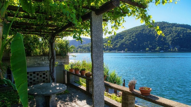 Une table et un banc en pierre, placés sous une pergola au bord d’un lac, avec des plantes en pot disposées sur une balustrade.