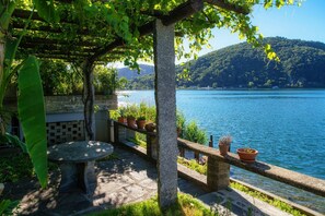 Une table et un banc en pierre, placés sous une pergola au bord d’un lac, avec des plantes en pot disposées sur une balustrade.