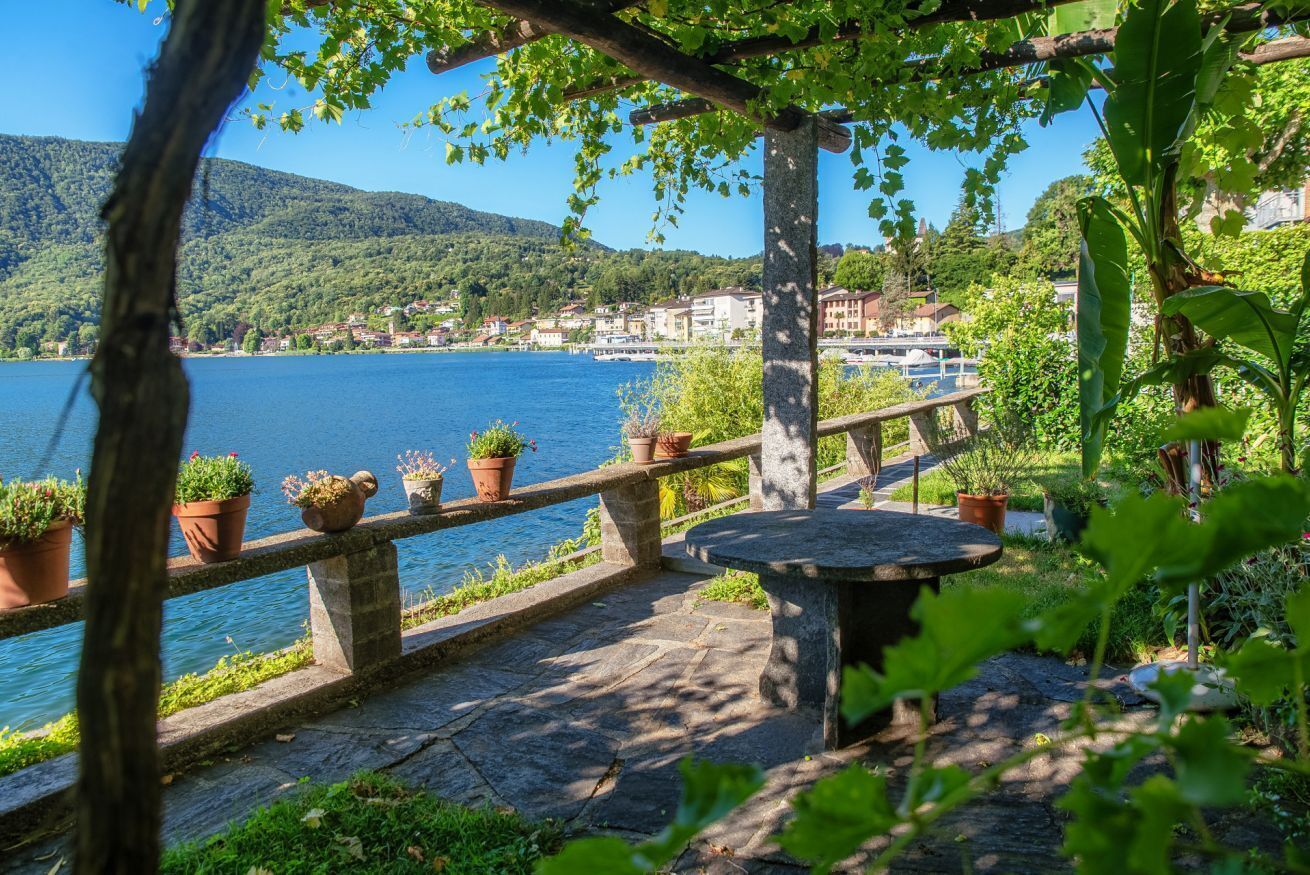 Ein Steinweg führt zu einer Sitzgelegenheit mit Blumentöpfen, mit Blick auf einen See und Gebäude im Hintergrund.
