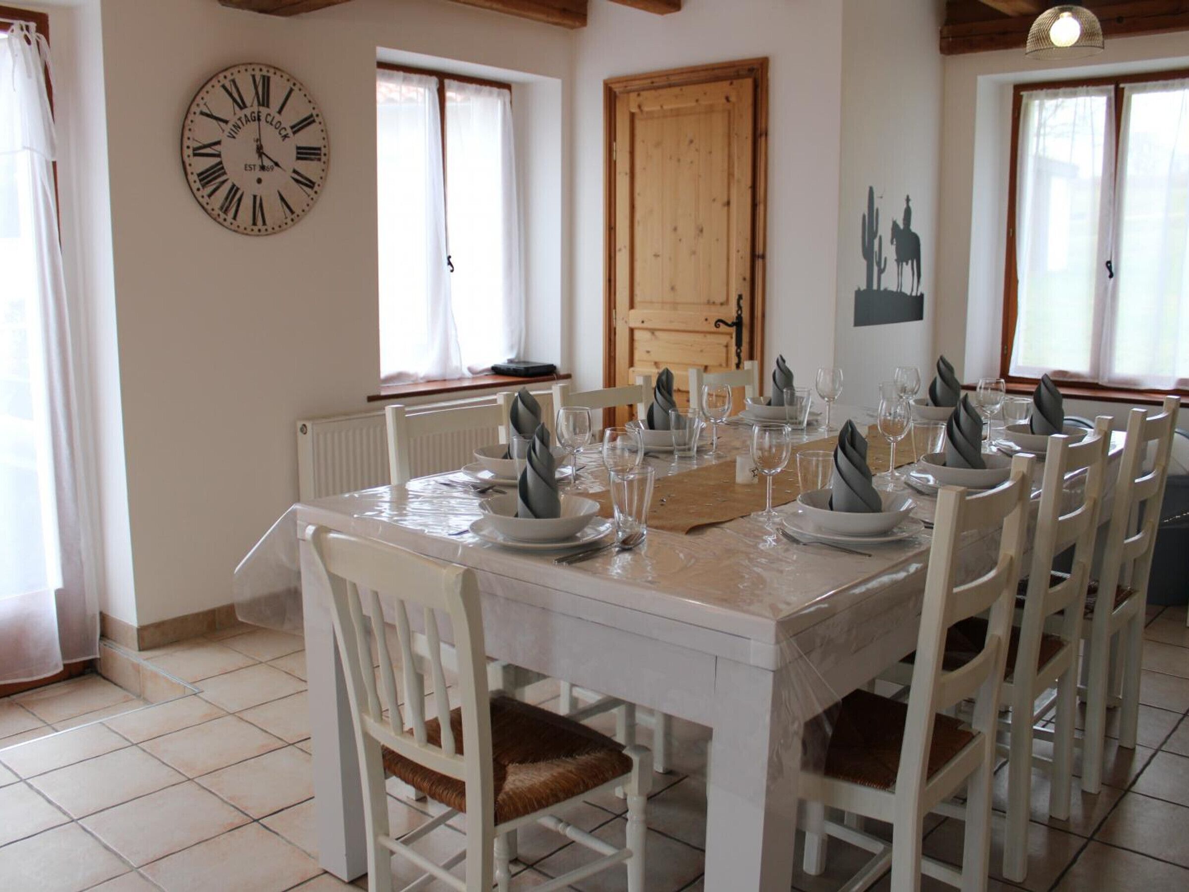 Een eetkamer met een tafel gedekt voor zes personen, witte stoelen, een antieke klok en een houten deur.