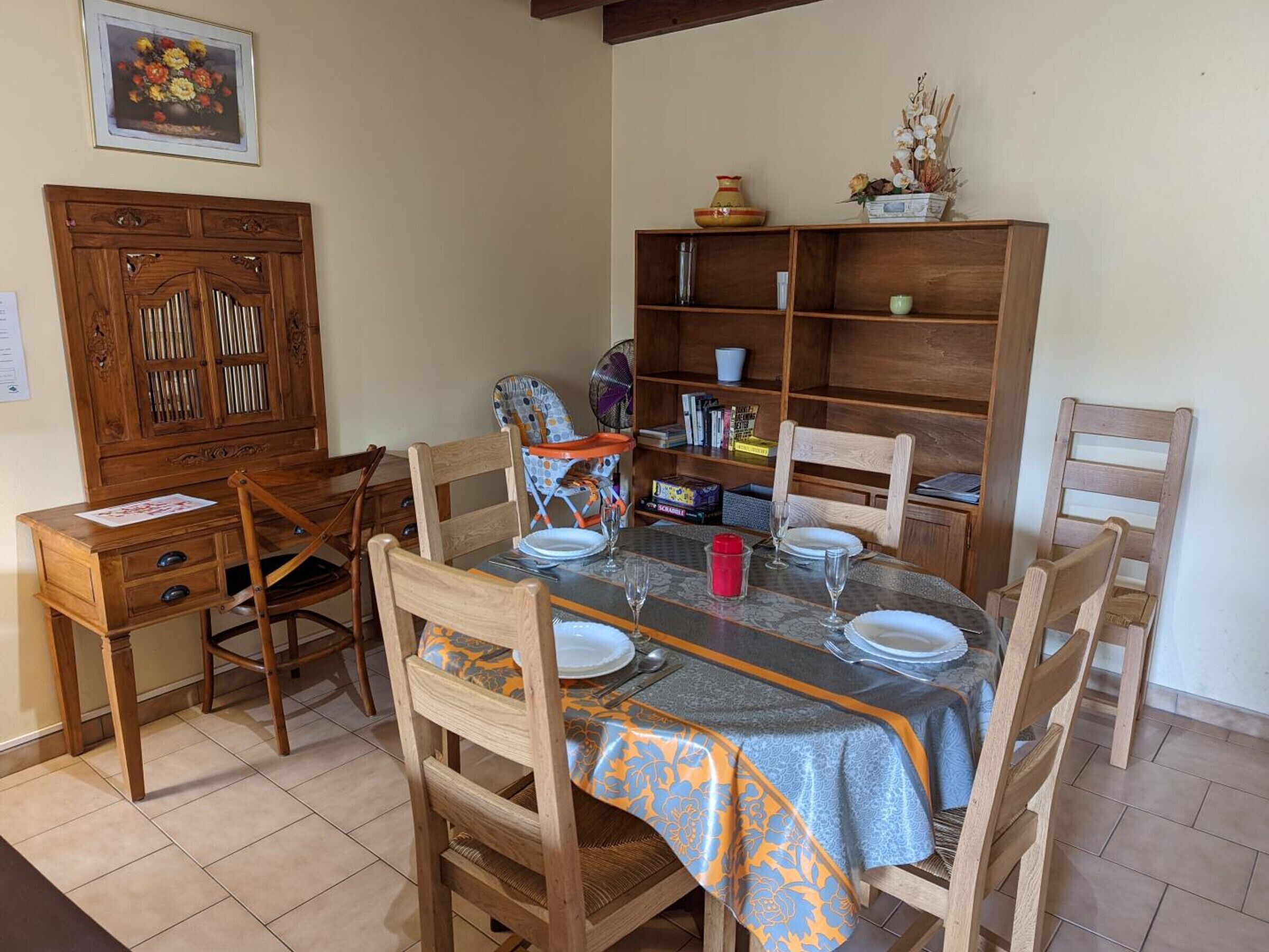 Een eetruimte met een houten tafel voor vier personen, omringd door houten stoelen. Links bevindt zich een houten kast met glazen deuren, en rechts een boekenkast met diverse spullen.