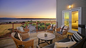 En terrass med möbler i rotting, ett runt bord och utsikt över havet vid solnedgången.