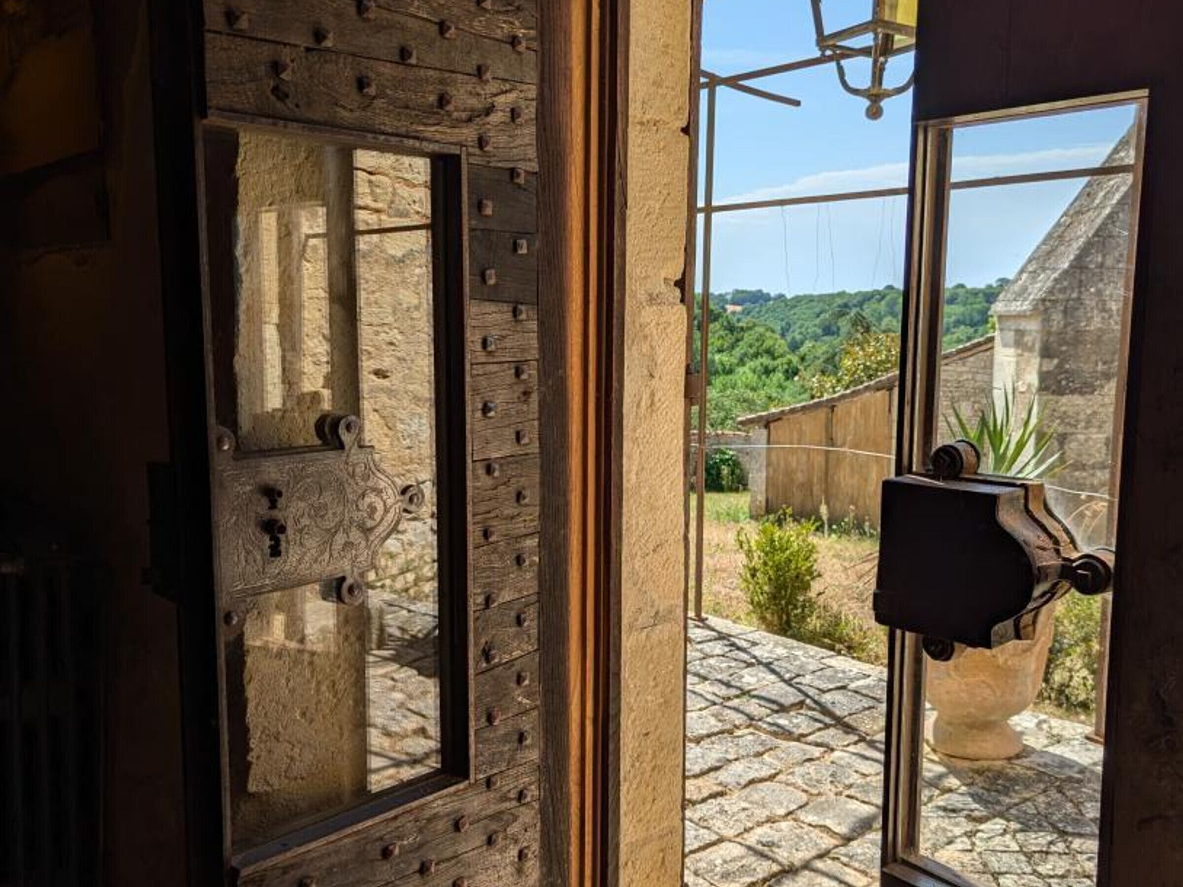 Una puerta de madera rústica con una ventana de piedra, que da a un patio empedrado con vistas a vegetación y un edificio a lo lejos.
