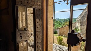Une porte en bois rustique, ornée d’une fenêtre en pierre, donne sur une cour dallée de pierres, d’où l’on aperçoit de la verdure et un bâtiment au loin.