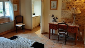 A room with a wooden floor, a desk with two chairs, a bed with a white bedspread, a window with curtains, and a stone wall with a lamp. - La Queue Rousse Cottage - Historic Home with Fireplace and Enclosed Garden (Vitrac-Saint-Vincent)