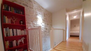 A hallway with a wooden floor, a red bookshelf filled with books, a white railing, and a door leading to another room.