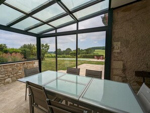 A conservatory with a glass table and chairs, stone walls, and a view of a garden and pool. - Charming Cottage with Heated Pool, Terrace, and Enclosed Garden in Aubeville (Val des Vignes)