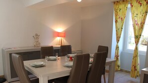 A dining area with a white table, six brown chairs, a red table lamp, and a window with yellow floral curtains. - Family cottage in village, pets allowed (Salles-Lavalette)