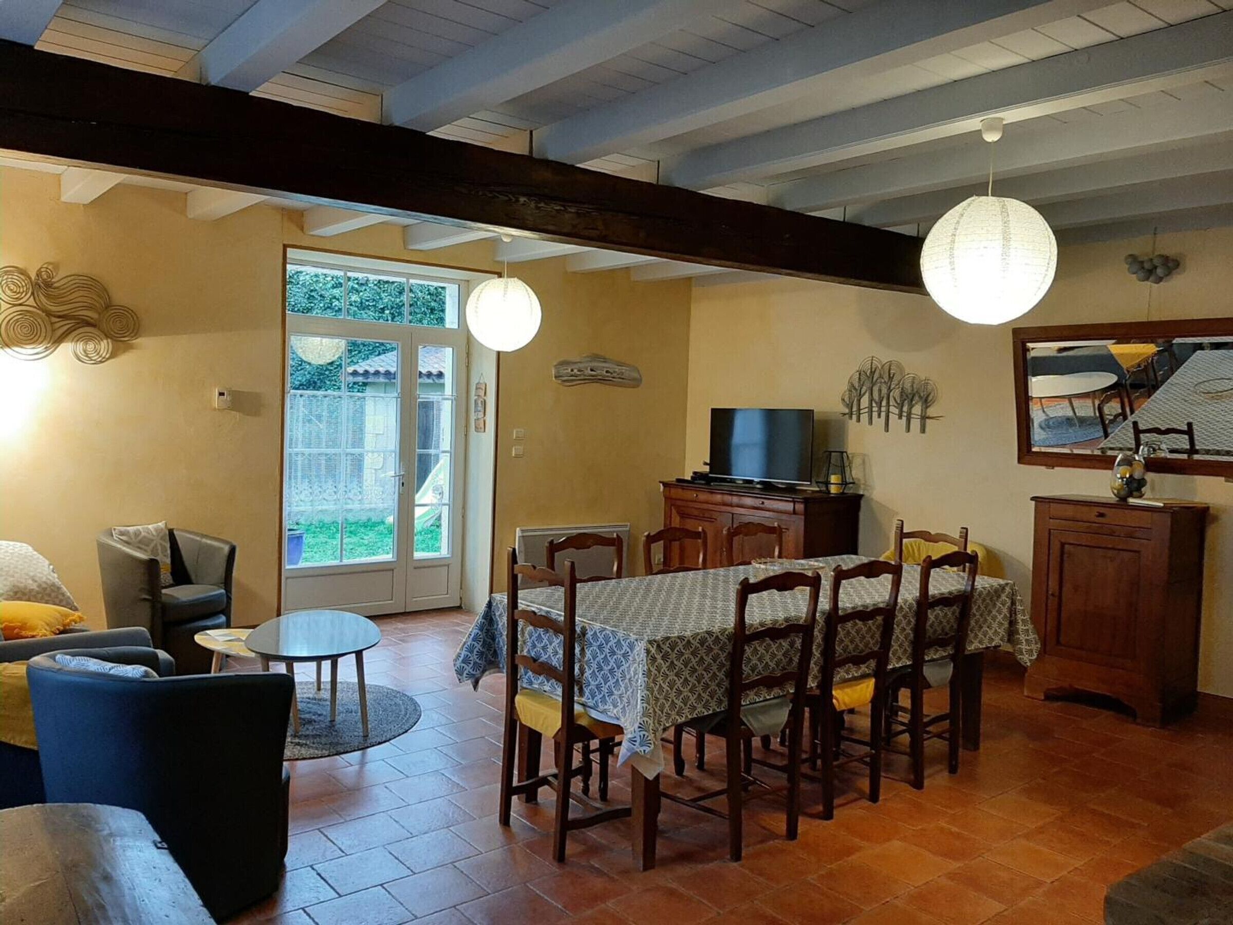 Un coin repas avec une table dressée pour six personnes, entourée de chaises en bois. Un téléviseur est posé sur un meuble en bois, et deux lampes suspendues se trouvent au-dessus de la table.