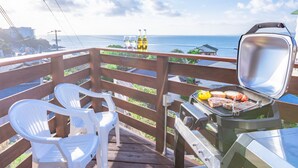 Un balcon avec un barbecue, deux chaises en plastique blanches et une vue sur l’océan.