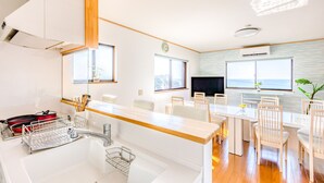 Une cuisine moderne avec un plan de travail en bois, des meubles blancs et un évier encastré. On y trouve un coin repas avec des chaises et une table blanches, ainsi qu’une grande fenêtre offrant une vue sur l’océan.