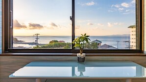Une vue depuis une fenêtre, avec une plante en pot sur une table en verre, donnant sur une zone côtière avec des bâtiments et une étendue d’eau.