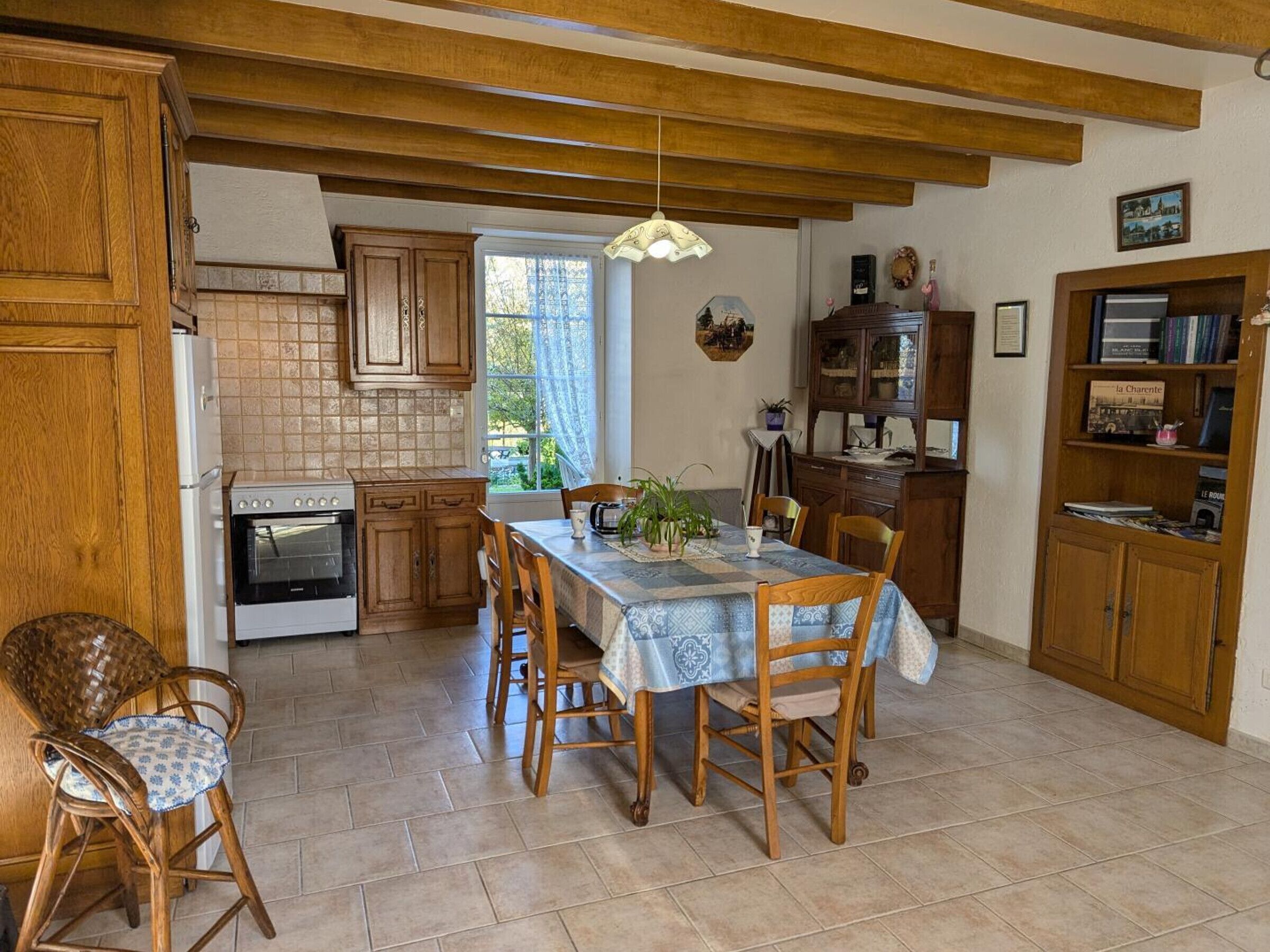 A kitchen with wooden cabinets, a dining table with chairs, a hanging light fixture, and a view of the outdoors through a window.