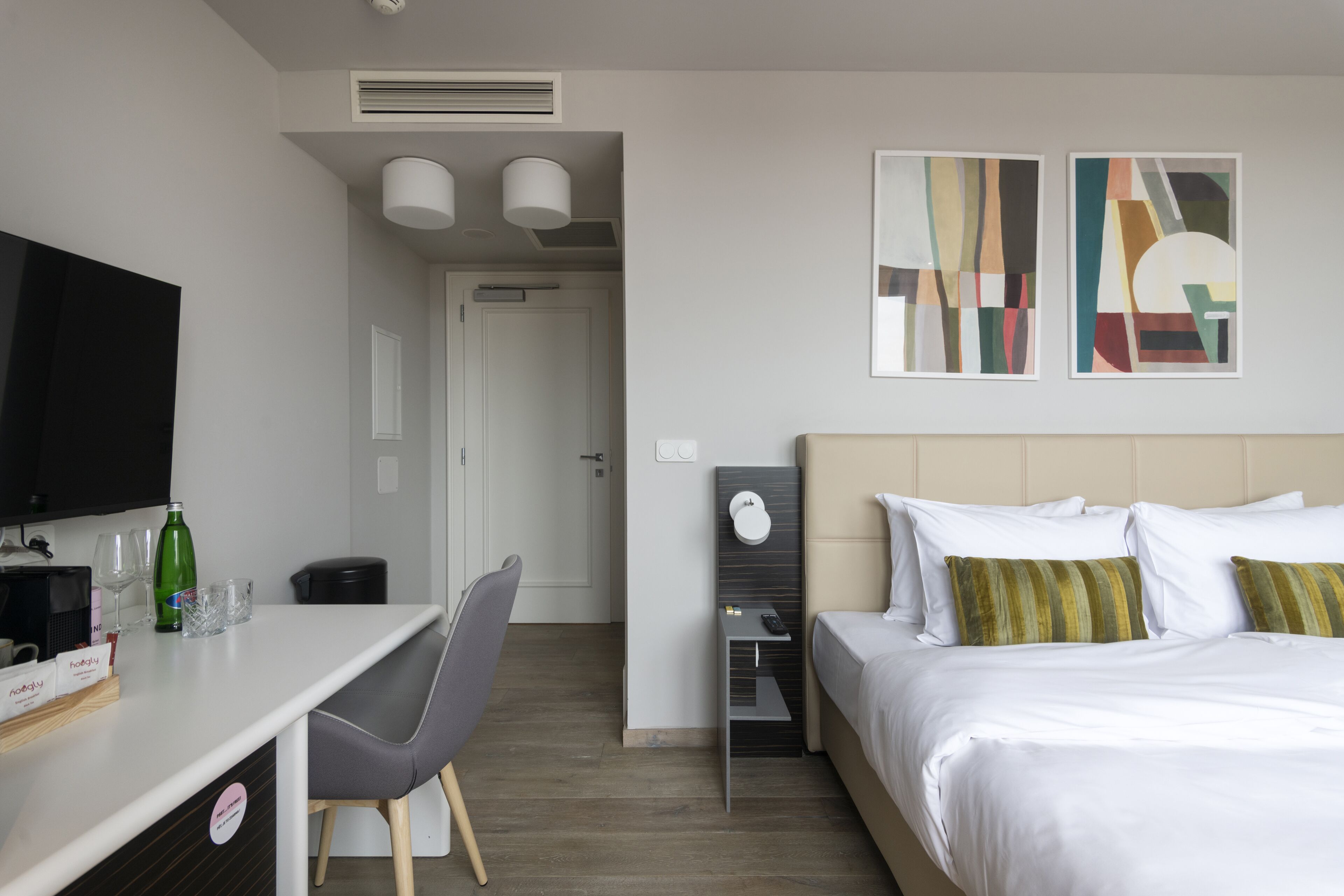 A modern hotel room with a bed, a desk, a chair, a TV, and artwork on the wall.