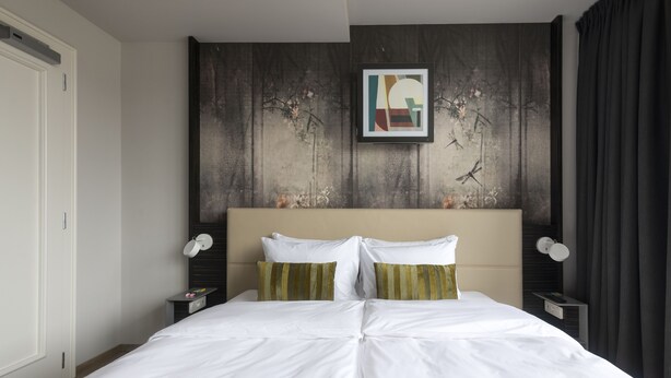 A hotel room with a large bed, a headboard with a framed artwork, a nightstand with a lamp, and a dark curtain. - Numa Prague Poise (Prague)