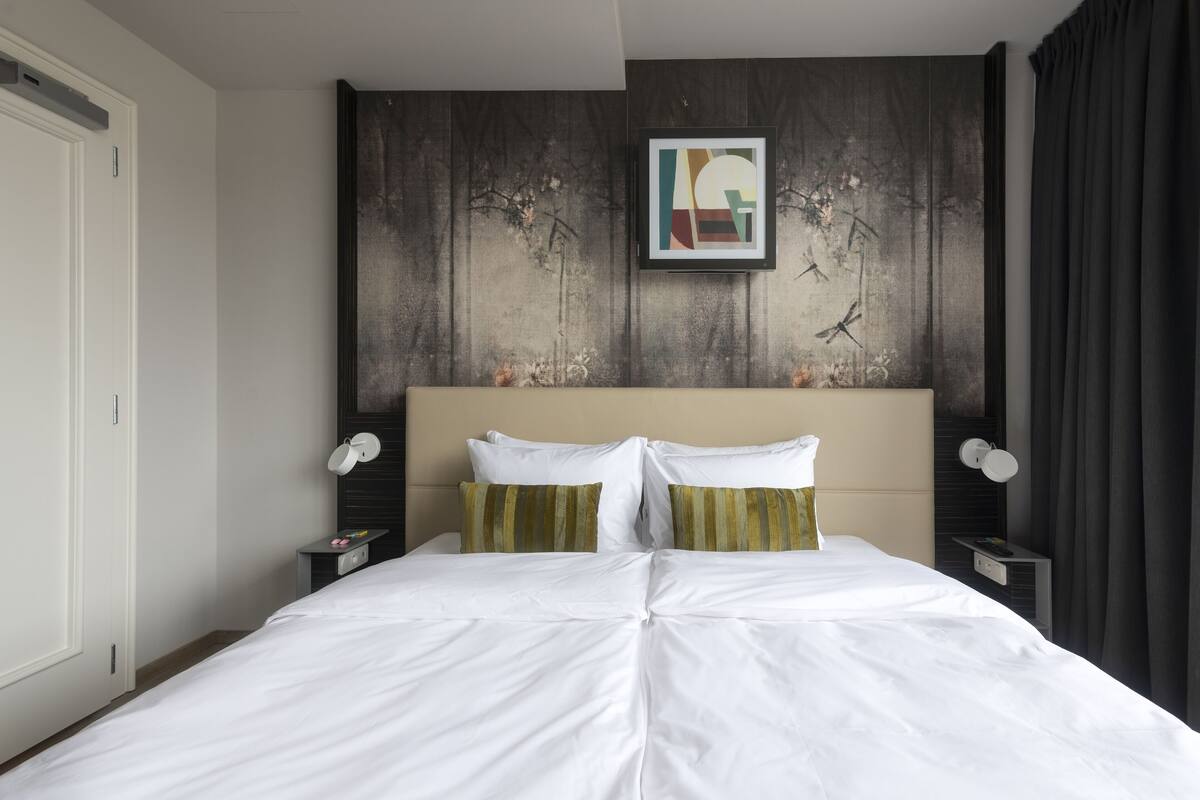 Une chambre d’hôtel avec un grand lit, une tête de lit ornée d’une œuvre d’art encadrée, une table de chevet avec une lampe et un rideau foncé.