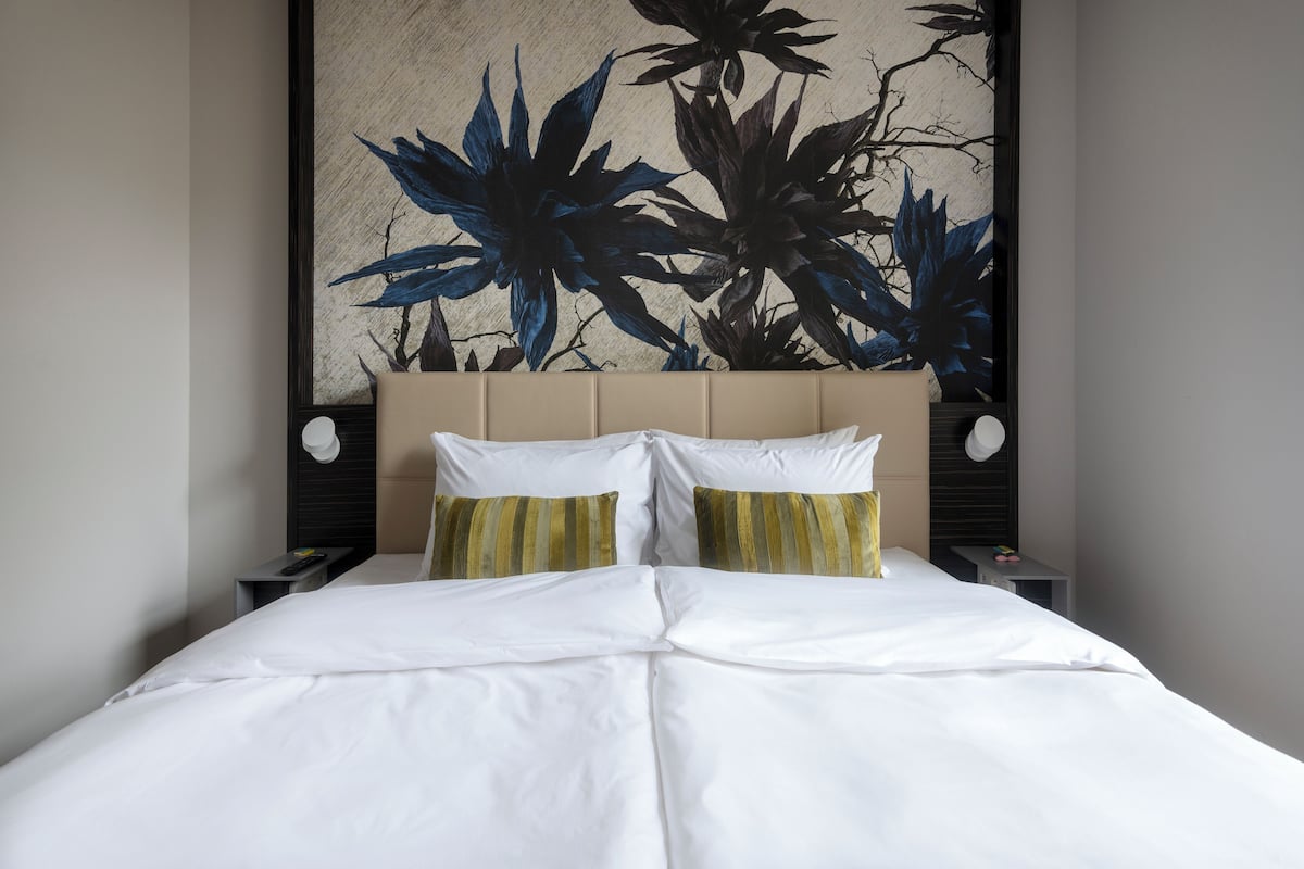 Un lit bien fait, avec du linge de lit blanc et deux oreillers, une tête de lit beige et une œuvre d’art fixée au mur représentant des feuillages bleus et verts.