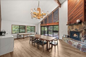 A spacious room with a large wooden dining table, a stone fireplace, and large windows offering a view of the outdoors.