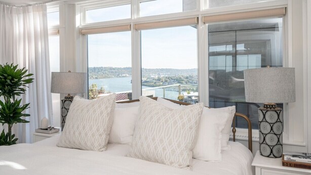 A bedroom with a large bed, two bedside tables, a potted plant, and a view of a body of water through large windows.