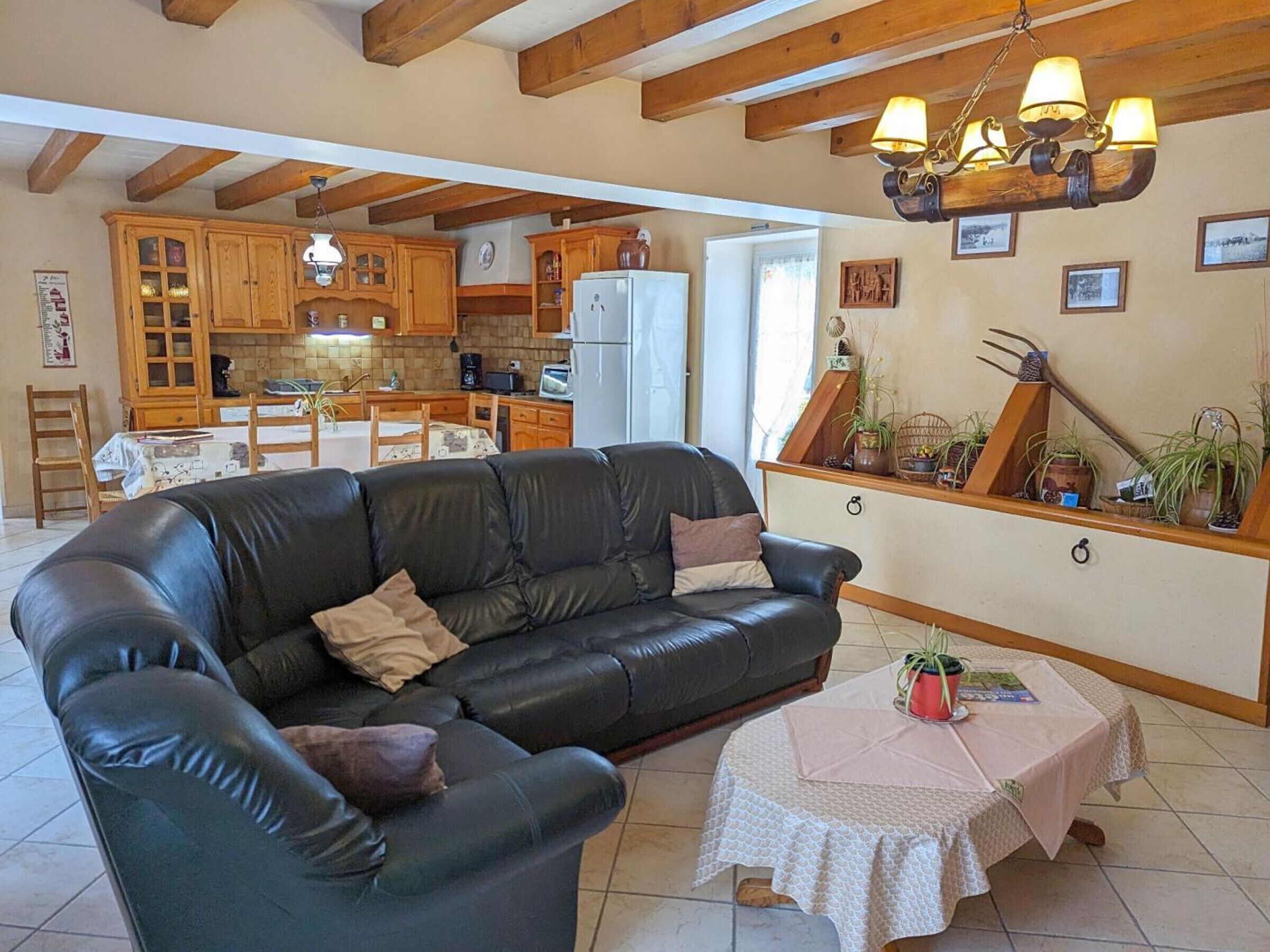 A living room with a black leather sofa, a small table with a white tablecloth, wooden cabinets, and a kitchen area with wooden beams.