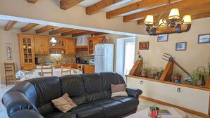 Una sala de estar con un sofá de cuero negro, una mesita con mantel blanco, armarios de madera y una zona de cocina con vigas de madera.