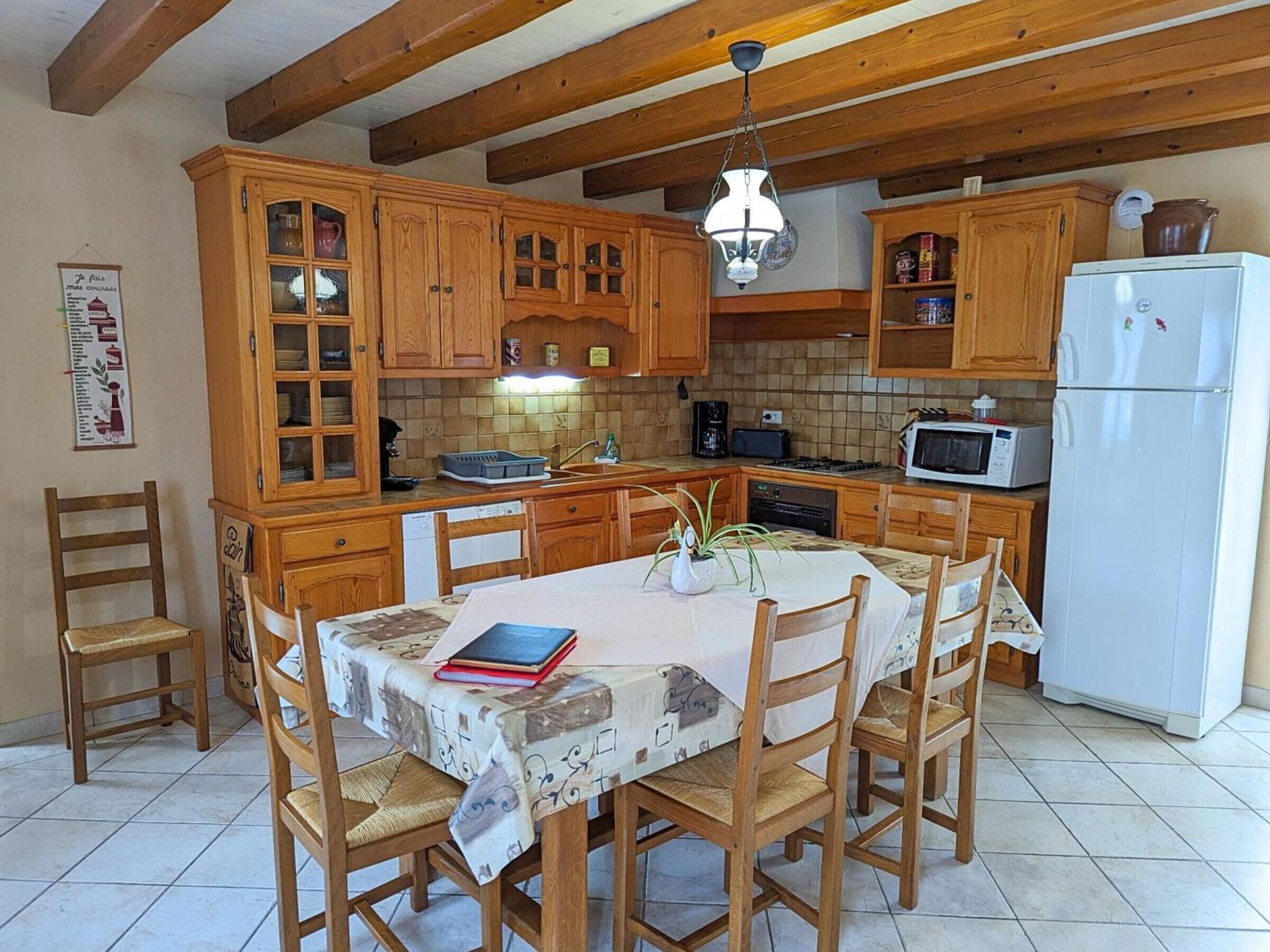 A kitchen with wooden cabinets, a dining table with chairs, a refrigerator, and a hanging light fixture.