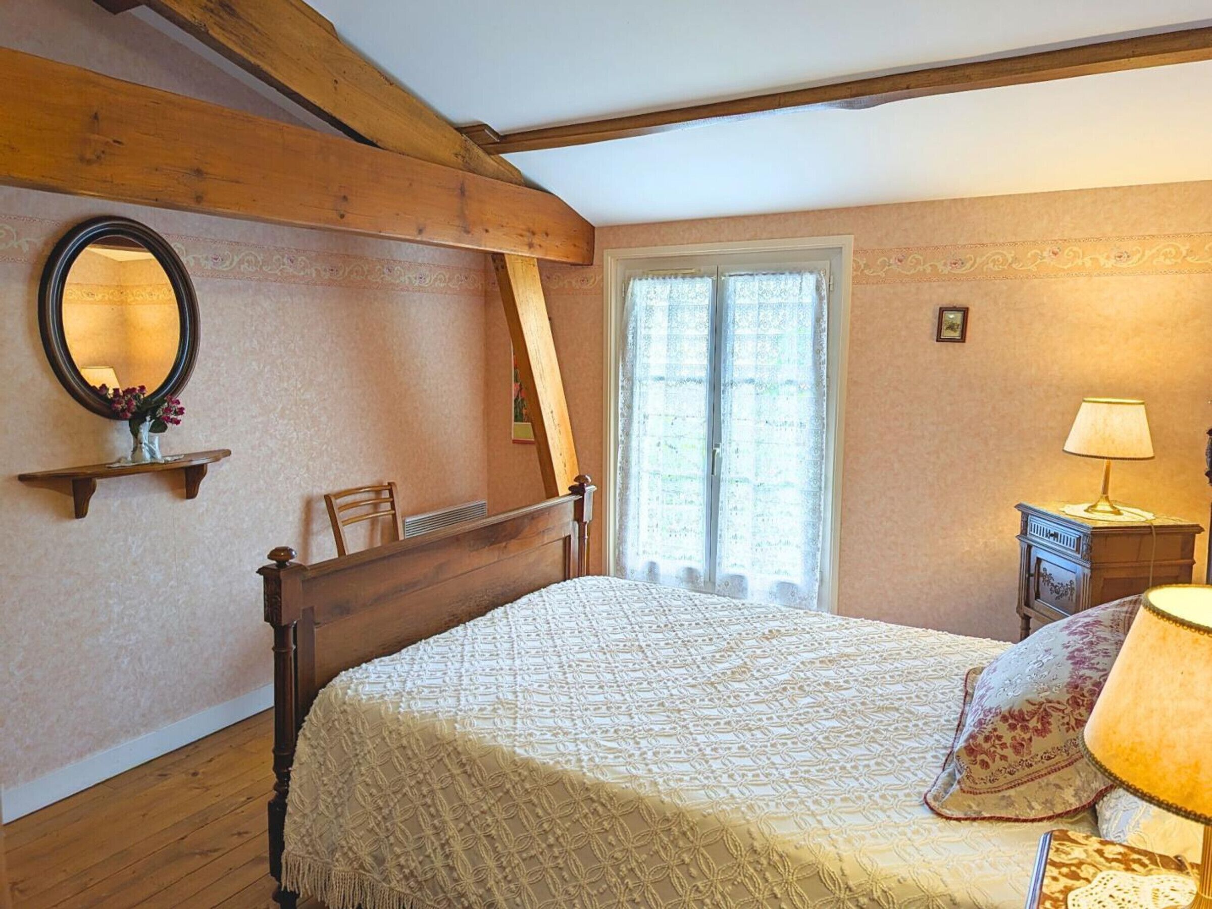 A bedroom with a wooden bed, a bedside table with a lamp, a window with curtains, and a mirror on the wall.