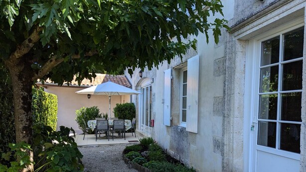 A courtyard with a table and chairs under a large umbrella, surrounded by greenery and a gravel path. - Charming Cottage with Pool, Fireplace, and Garden in Vineyard Estate (Saint-Cybardeaux)