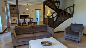 A living room with a sofa, armchair, and coffee table, a staircase, and a dining area with a table and chairs. - Charming Cottage with Pool, Fireplace, and Garden in Vineyard Estate (Saint-Cybardeaux)