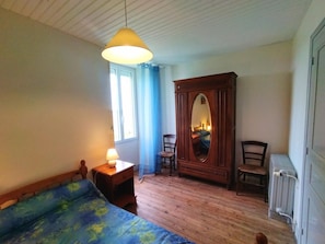 A bedroom with a bed, a wooden wardrobe, a chair, a bedside table with a lamp, and a radiator.