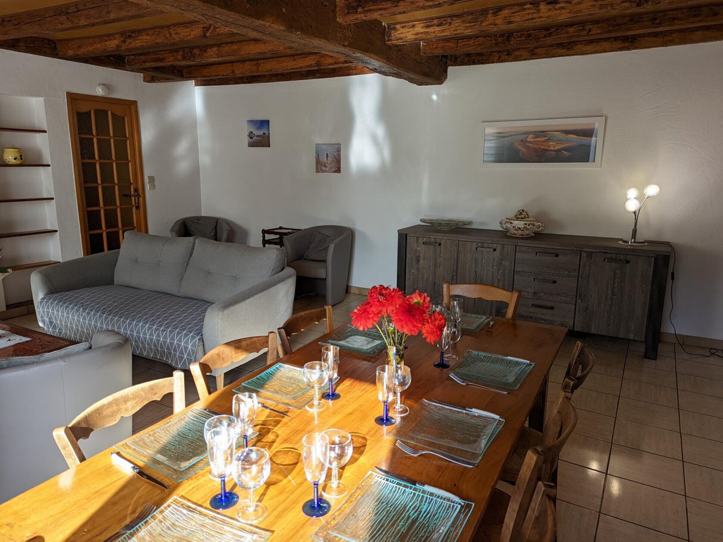 A dining area with a wooden table set for six, surrounded by chairs. There is a sofa and a cabinet in the background.