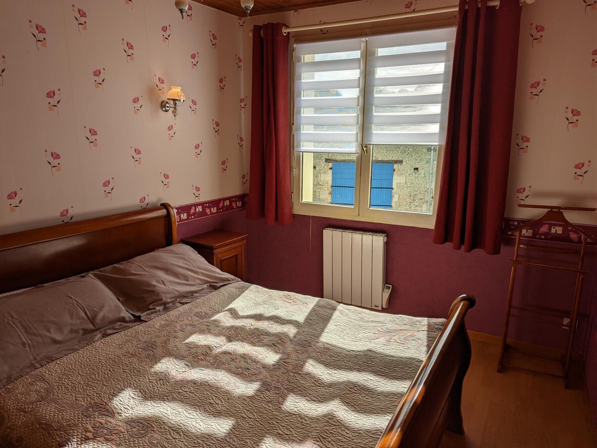 A bedroom with a bed, a nightstand, a radiator, a window with curtains, and a wall with floral wallpaper.