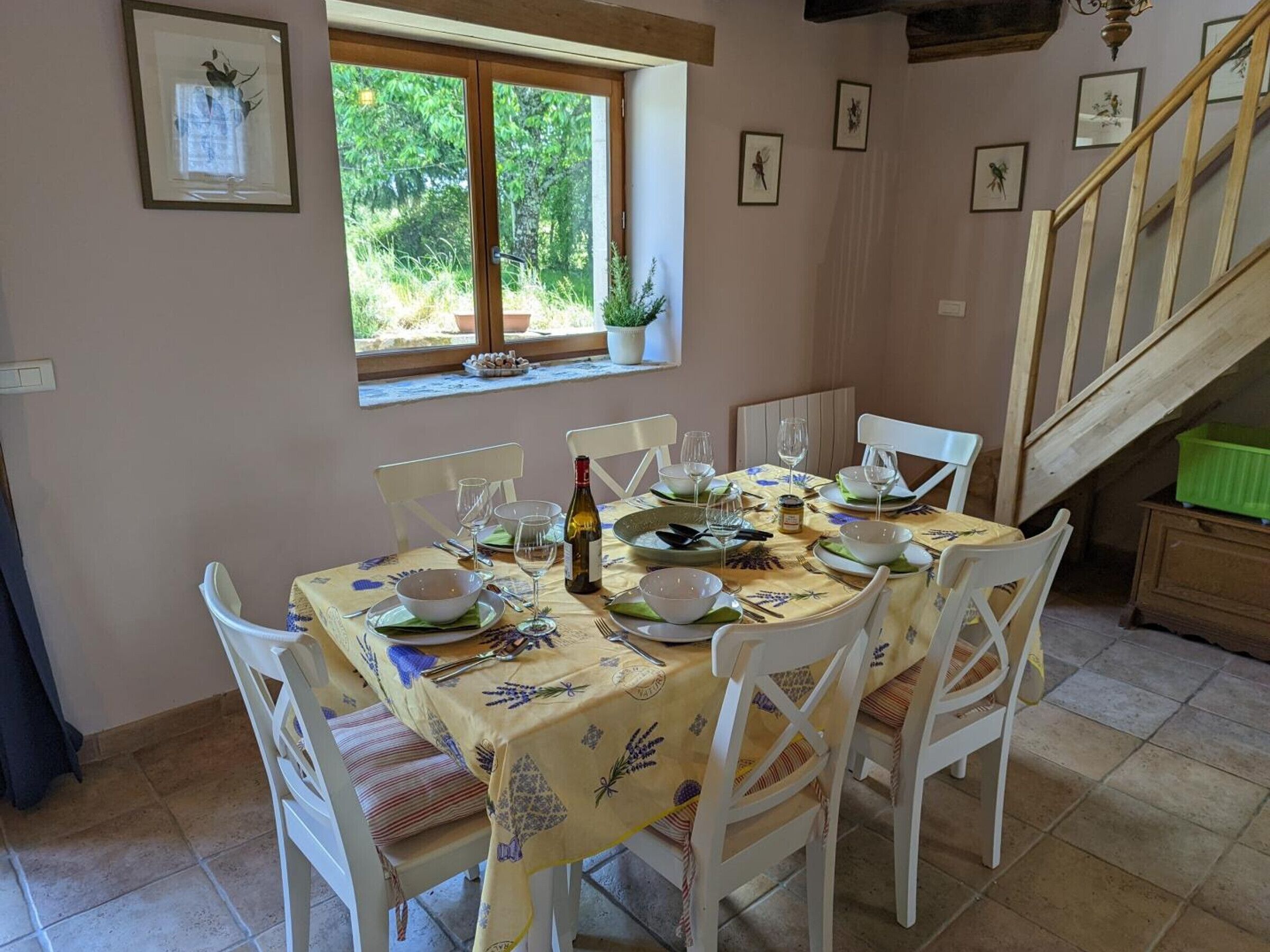Un coin repas avec une table dressée pour six personnes, des chaises blanches, une fenêtre donnant sur un paysage verdoyant et un escalier.