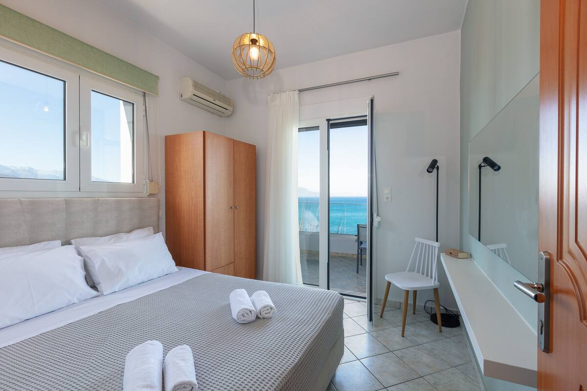 A bedroom with a bed, a wooden wardrobe, a bathroom with a mirror, and a view of the sea.