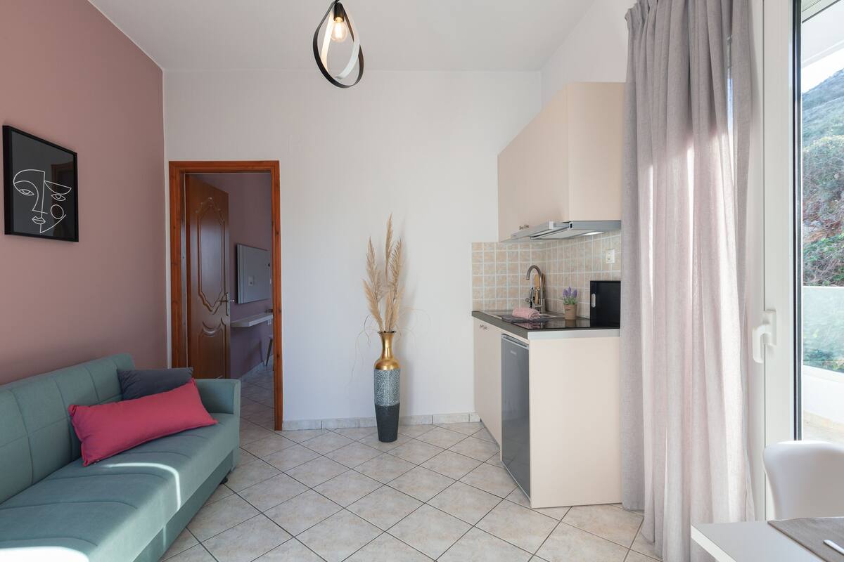 A compact living space with a kitchenette, a sofa with cushions, a vase with dried plants, and a window with curtains.